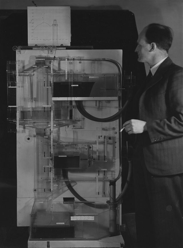 Bill Phillips stands beside his machine of clear tanks and pipes. A pen on top has drawn a curve on graph paper.